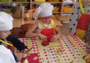 Dwoje dzieci przy stoliku kroi jabłka nożem.